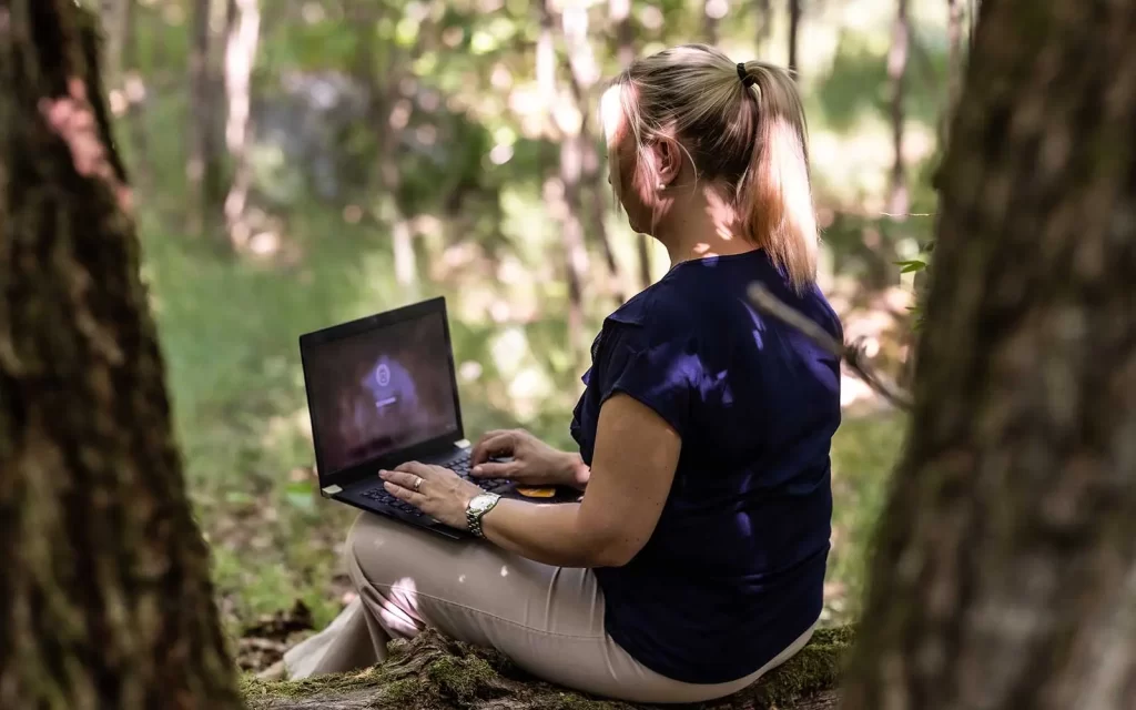 kvinna sitter i skogen på ett träd med sin laptop i knät