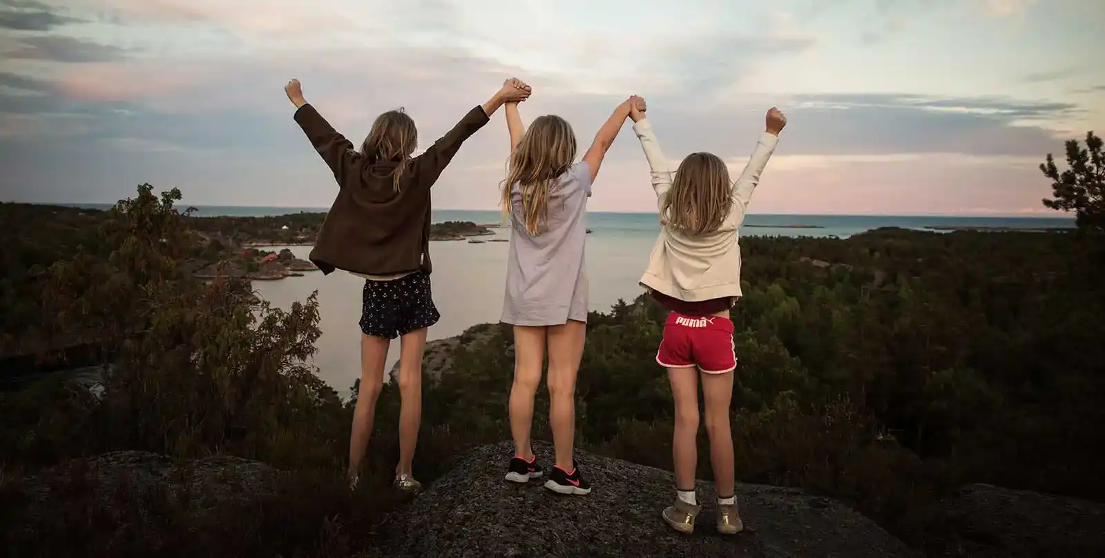 Tre glada barn som står med händerna up i vädret med ryggen mot kameran